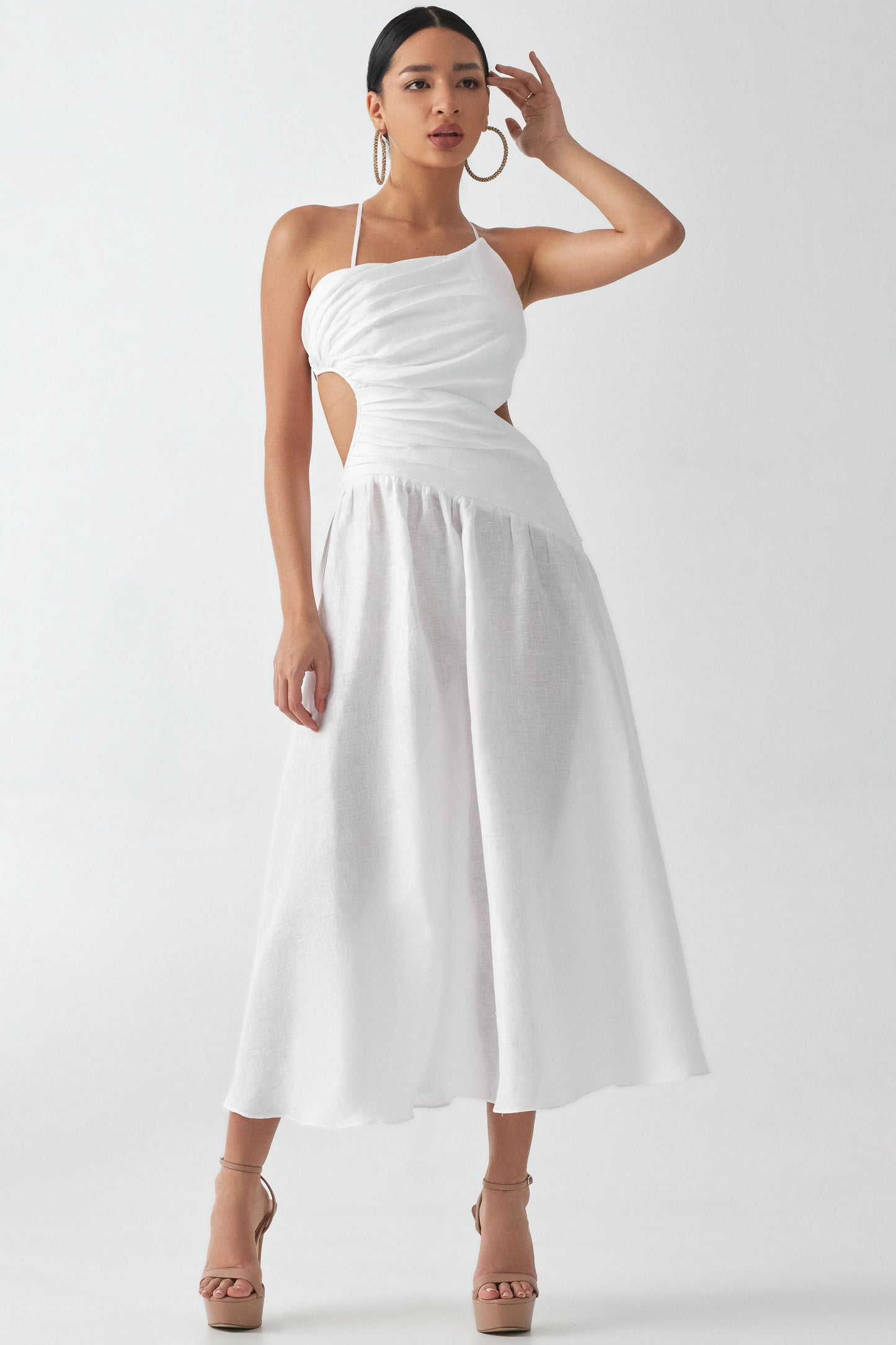 A model wearing a white maxi dress with ruched front and open-side design, and a draping waist seam. The dress has an open back and is paired with tan platform heels.
